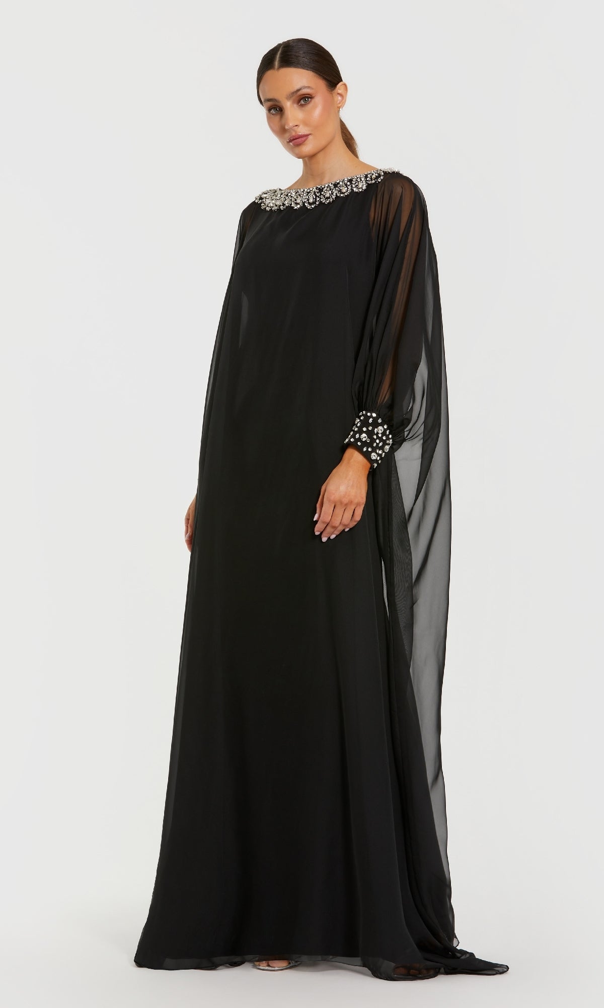 A woman models the Mac Duggal 56203 long formal dress, a black chiffon gown with rhinestone embellishments, sheer sleeves, and detailed neckline and cuffs, set against a plain white background.